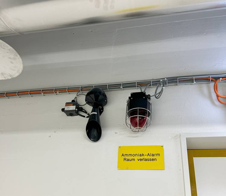Ammoniakwarnanlage mit akustischem Signalgeber und roter Warnleuchte über gelbem Hinweisschild, montiert an einer weißen Wand in einem Technikraum.