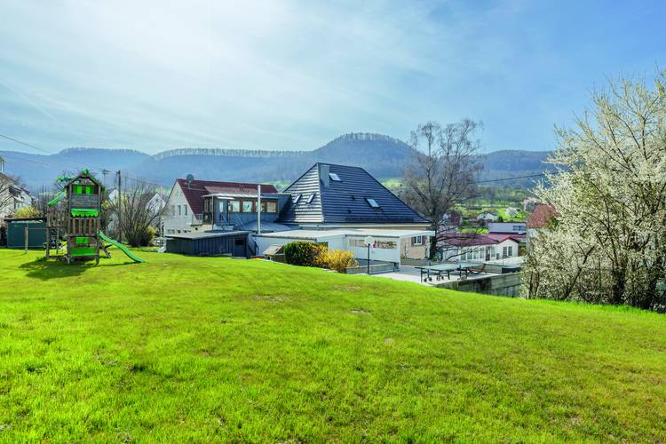 Mehrere moderne Wohngebäude auf begrüntem Gelände, Spielplatz im Vordergrund, umgeben von Bäumen und Hügeln - Fokus auf Gebäudetechnik-Infrastruktur.