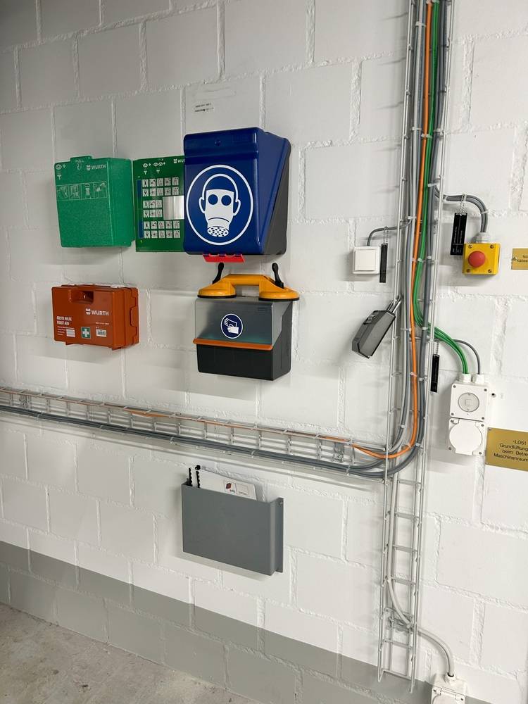 Wandmontierte Notfallausrüstung mit Erste-Hilfe-Kästen, PSA-Hinweisschild sowie Elektroinstallationen in einem Gebäudetechnikbereich.