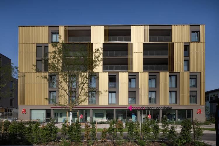 Modernes viergeschossiges Gebäude mit goldenen Fassadenpaneelen, Balkonen und integrierter Apotheke - Beispiel energieeffizienter Gebäudetechnik.
