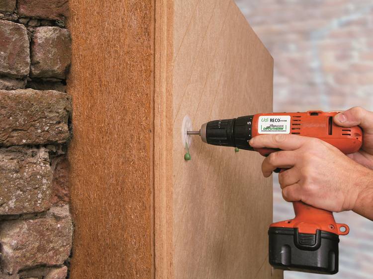 Nahaufnahme: Handwerker bohrt mit orangefarbenem Akkuschrauber Loch in Holzplatte an gemauerter Wand, typische Arbeit in der Gebäudetechnik.