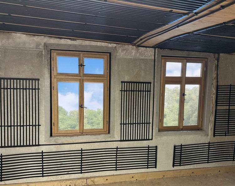 Zwei Holzrahmenfenster in einer Betonwand, darunter und seitlich schwarze Heiz- oder Lüftungspaneele - Beispiel für Fassadenintegration in der Gebäudetechnik.
