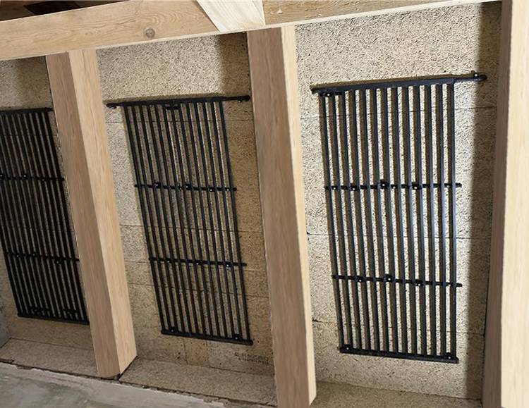 Drei senkrechte schwarze Metallgitter als Lüftungselemente auf einer Steinwand, eingefasst von hellen Holzträgern, Beispiel technischer Gebäudeausstattung.