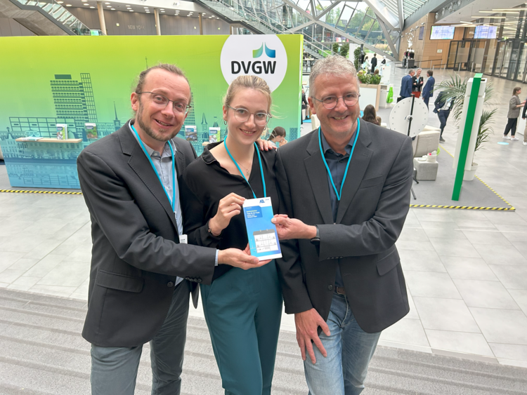 Drei Fachleute präsentieren gemeinsam eine DVGW-Broschüre auf einer Branchenveranstaltung für Gebäudetechnik in modernem Konferenzraum.
