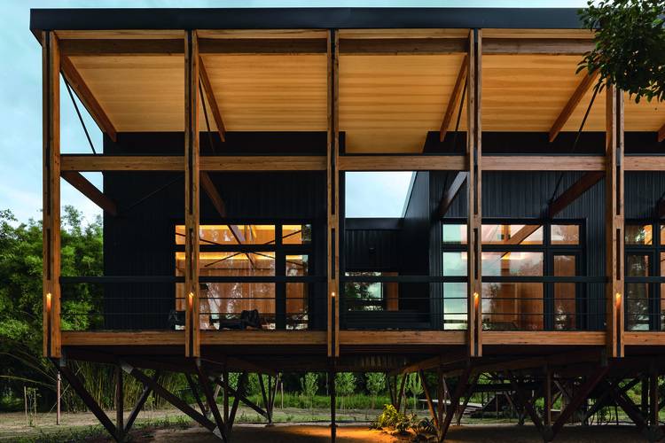 Energieeffizientes Wohngebäude auf Stelzen, großflächige Verglasung und Holzträger, moderne Fassadentechnik, beleuchtet bei Abenddämmerung.