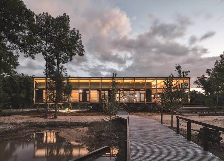 Modernes Bürogebäude mit verglaster Fassade auf Stelzen, energieeffizient beleuchtet, Holzsteg über Wasser, umgeben von Bäumen bei Dämmerung.