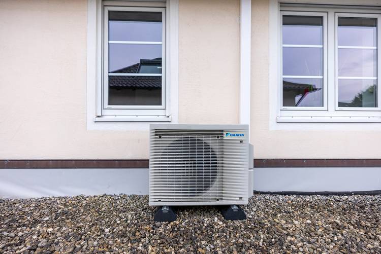 Außen-Klimagerät auf Kiesfläche unter zwei Fenstern an einer beigen Fassade, Beispiel für Split-Klimaanlage in der Gebäudetechnik.