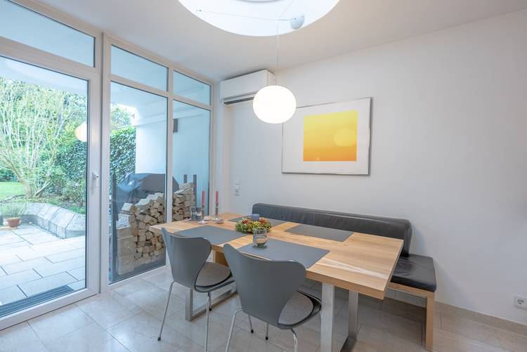 Moderner Essbereich mit Holztisch, zwei Stühlen und Bank vor großer Fensterfront; Ausblick auf Terrasse mit gestapeltem Brennholz, Fokus Gebäudehülle.