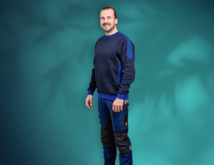 Gebäudetechniker im dunkelblauen Sweatshirt und blauer Arbeitshose vor türkisfarbenem Hintergrund, Symbolbild für Fachkräfte in der SHK-Branche.