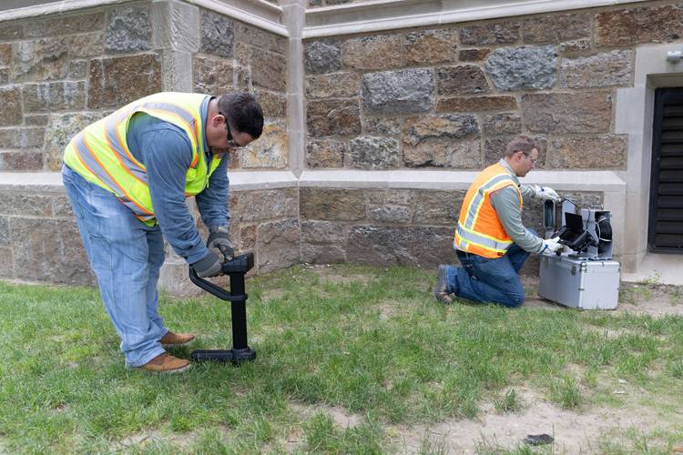 Zwei Fachkräfte in Warnwesten entnehmen mit Bodenproben-Gerät neben einem Bestandsgebäude Bodenproben zur Untersuchung für Gebäudeplanung und -sanierung.