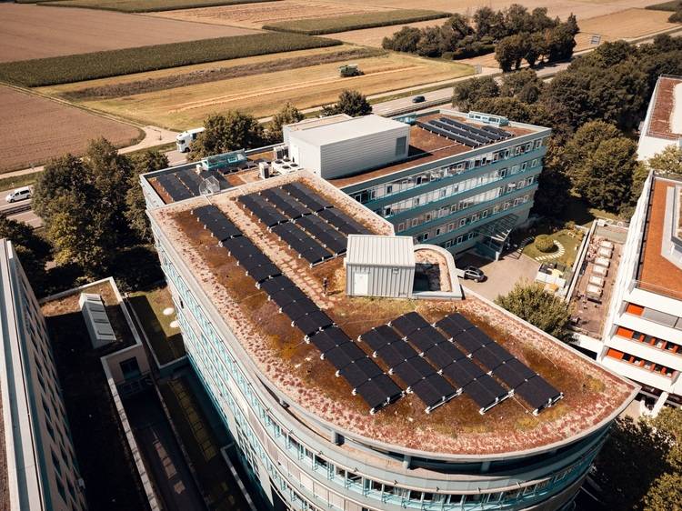 Drohnenaufnahme: Modernes Gebäude mit extensiver Dachbegrünung und Photovoltaikanlage, umgeben von Grünflächen - Fokus auf Energieeffizienz und nachhaltige Gebäudehülle.