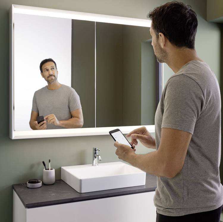 Fachhandwerker prüft Sanitärinstallation im Badezimmer, dokumentiert Daten per Smartphone, Spiegel und Waschbecken im Hintergrund sichtbar.