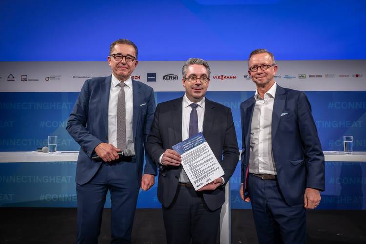 Drei Branchenvertreter in Anzügen auf Bühnenpodium, mittig wird eine Urkunde überreicht; blauer Hintergrund mit Gebäudetechnik-Eventbranding.