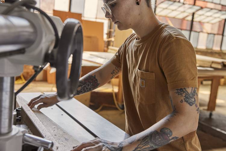 Fachkraft bedient Holzbearbeitungsmaschine in heller Werkstatt, trägt braunes T-Shirt und Schutzbrille - Symbolbild für handwerkliche Gebäudetechnik.