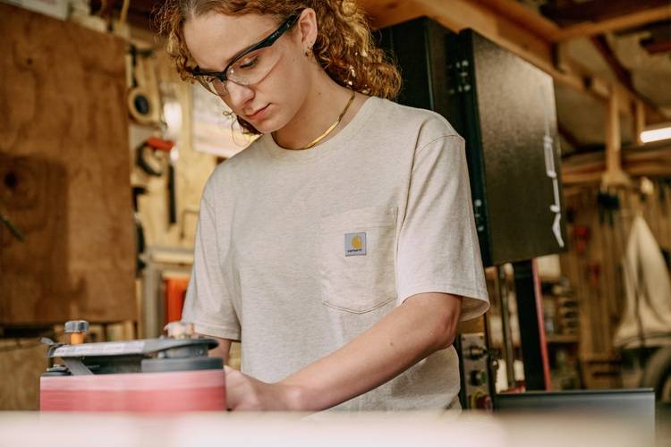 Fachkraft mit Schutzbrille schleift Holzbauteil in Werkstatt - Fokus auf sichere Verarbeitung und Oberflächenoptimierung für den Gebäudetechnikbereich.