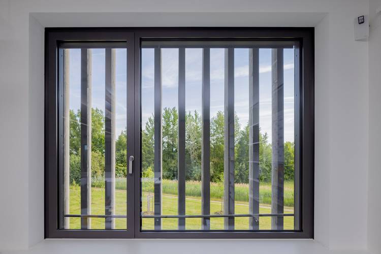 Großes Fenster mit vertikalen Metallstreben in einem modernen Gebäudetechnik-Objekt, Blick auf Außenanlage mit Grünfläche und Bäumen.