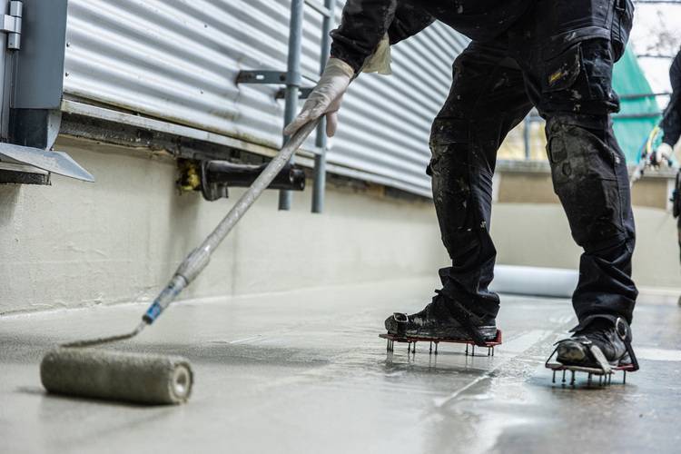 Fachhandwerker trägt mit Stachelsohlen und langem Roller eine Abdichtungsbeschichtung auf einem Boden im Neubau gemäß TGA-Standards auf.
