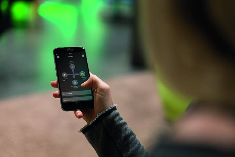 Fachkraft steuert Gebäudetechnik über Smartphone-App mit Bedienoberfläche, im Hintergrund unscharfe grüne Statusanzeigen für Energieeffizienz.