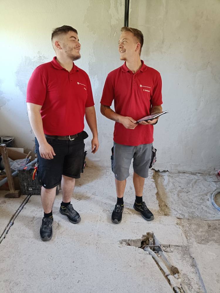 Zwei Fachhandwerker in roter Arbeitskleidung begutachten gut gelaunt den Baufortschritt bei der Haustechnik-Installation im sanierten Innenraum.
