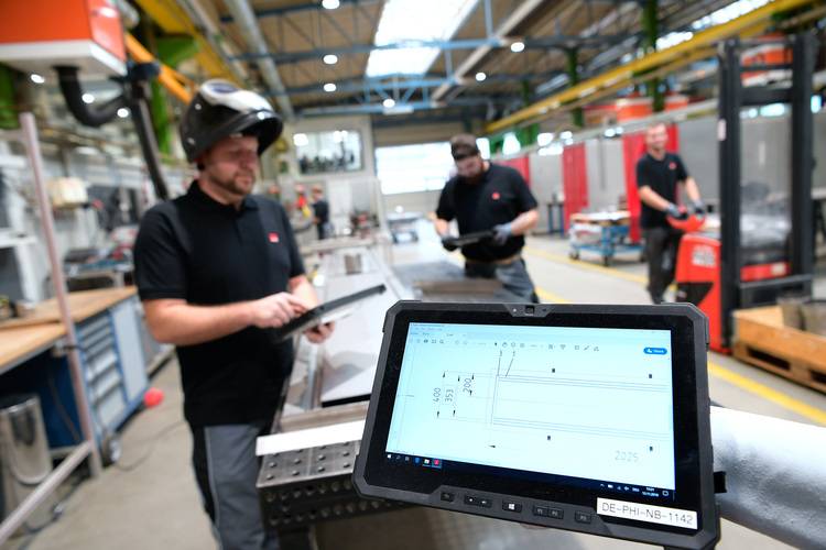 Tablet mit Baustellenplan zur Heizungsinstallation in Fabrikhalle; im Hintergrund arbeiten Fachkräfte der Gebäudetechnik mit Schutzkleidung.