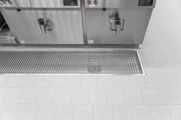 Bodenablauf aus Edelstahl mit Rost in gefliester Industrieumgebung, geeignet für hygienische und sichere Entwässerung in gewerblichen Küchen.