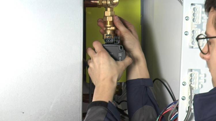 Fachhandwerker justiert ein Heizungsventil in einem Metallverteilerkasten; sichtbare Rohrleitungen und Elektroverkabelung für die Gebäudetechnik.