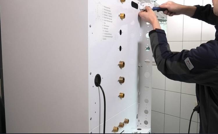 Fachkraft installiert weißes Steuerungsmodul für Gebäudetechnik an gefliester Wand; sichtbar sind mehrere Rohranschlüsse und Montagetool.