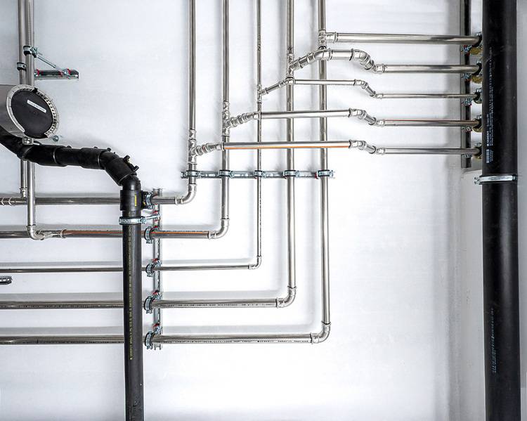 Installierte Heizungsrohrleitungen mit glänzenden Armaturen an weißer Wand, links eine schwarz gedämmte Leitung; Beispiel für TGA-Installation.