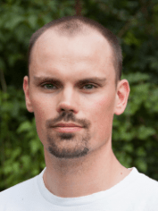 Ein Mann mit kurz braunen Haaren und Kinnbart im weißen Hemd steht vor Grünpflanzen, typisches Porträt für Gebäudetechnik-Experten auf haustec.de.