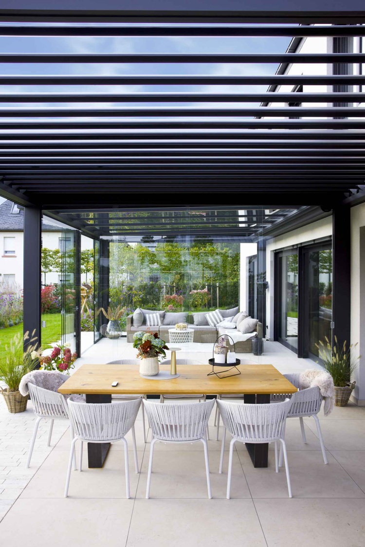 Überdachte Terrasse mit Holztisch, weißen Stühlen und Pflanzkübeln - Beispiel für moderne Außenraumgestaltung im Objektbau, Fokus Gebäudetechnik.