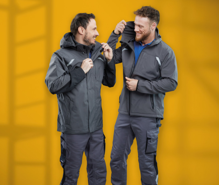 Zwei Fachhandwerker in grauer Arbeitskleidung schließen ihre Jacken, lächeln und stehen vor gelbem Hintergrund - Symbol für Gebäudetechnik-Teamwork.