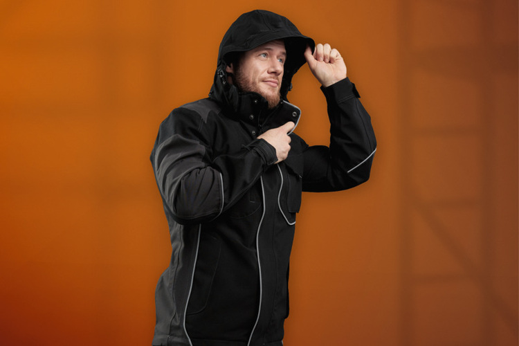 Fachhandwerker in schwarzer Arbeitsjacke hält Smartphone vor Brust, orangefarbener Hintergrund - Symbolbild für moderne Gebäudetechnik-Kommunikation.