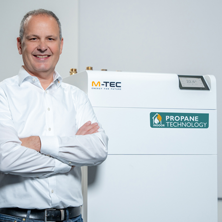 Fachmann in weißem Hemd neben einer Propanwärmepumpe von M-TEC, präsentiert innovative Gebäudetechnik für effiziente Energieversorgung.