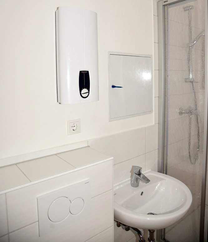 Kompaktes Bad mit wandhängendem Elektro-Durchlauferhitzer, Waschtisch, Einhebelmischer, barrierefreier Dusche und modernen Spültasten.