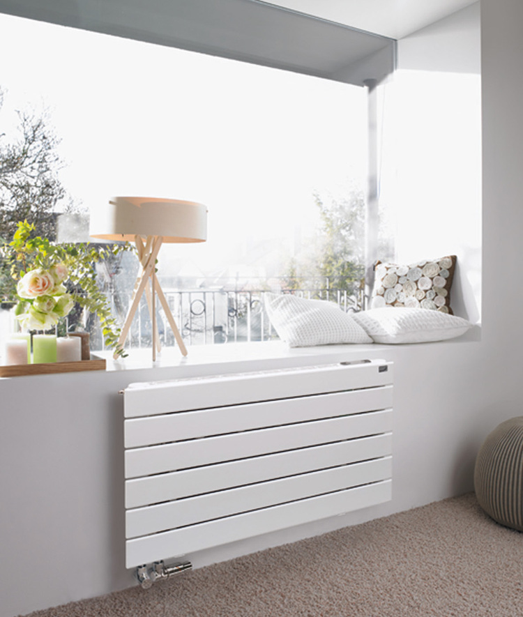 Moderner weißer Heizkörper unter einem Fensterplatz mit Sitzkissen; Beispiel für effiziente Wärmeabgabe in lichtdurchfluteten Gebäuden.