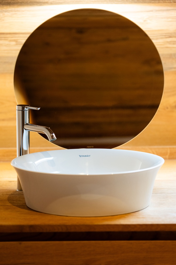 Waschtischanlage mit moderner weißer Keramik, verchromtem Einhebelmischer und rundem Spiegel auf Holzkonsole - Beispiel für zeitgemäße Sanitärinstallation.