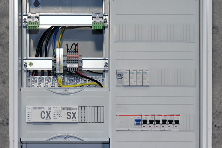 Schaltschrank mit Leistungsschutzschaltern, Verdrahtung und beschrifteten Schaltern, montiert an grauer Wand in einem modernen Gebäudetechnik-Umfeld.