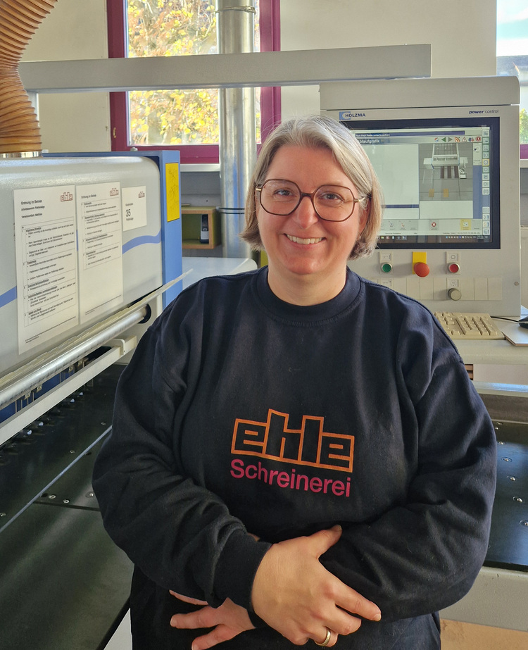 Fachkraft mit Brille im "Schreinerei"-Sweatshirt steht in Werkstatt vor Holzbearbeitungsmaschinen, thematisch Gebäudetechnik-Umfeld.