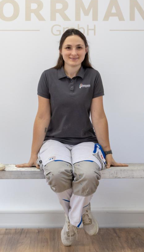 Fachfrau in Arbeitskleidung sitzt lächelnd auf einer Bank vor weißer Wand mit Logo - Symbol für Kompetenz in der Gebäudetechnikbranche.