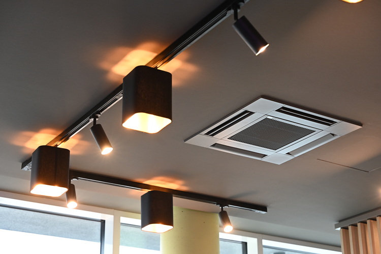 Abgehängte Decke mit modularen LED-Lichtschienen und quadratischem Lüftungsventil, Montage nahe bodentiefer Verglasung in einem modernen Bürogebäude.