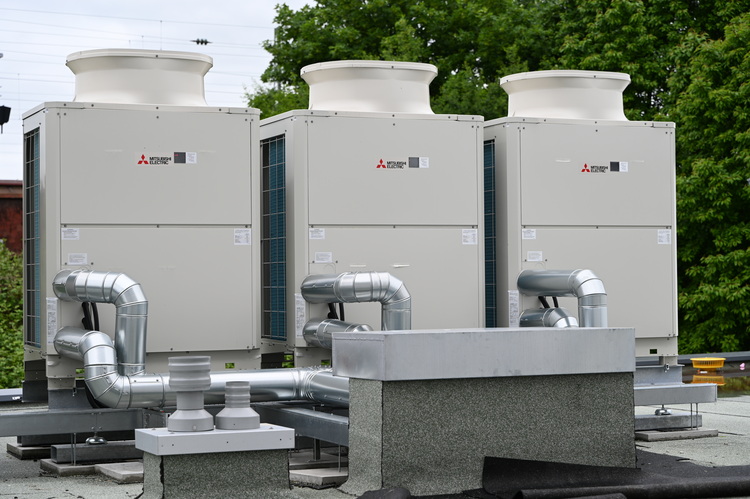 Drei große industrielle Klimageräte mit Metallkanälen auf einem Flachdach, typische Installation für moderne Gebäudetechnik, Bäume im Hintergrund.