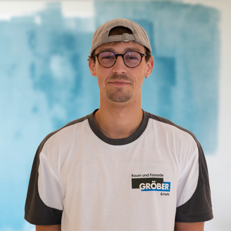 Junger Mitarbeiter der Firma Gröber trägt Firmen-T-Shirt und Kappe, steht vor blauem Hintergrund - Fokus auf Gebäudetechnik-Branche.
