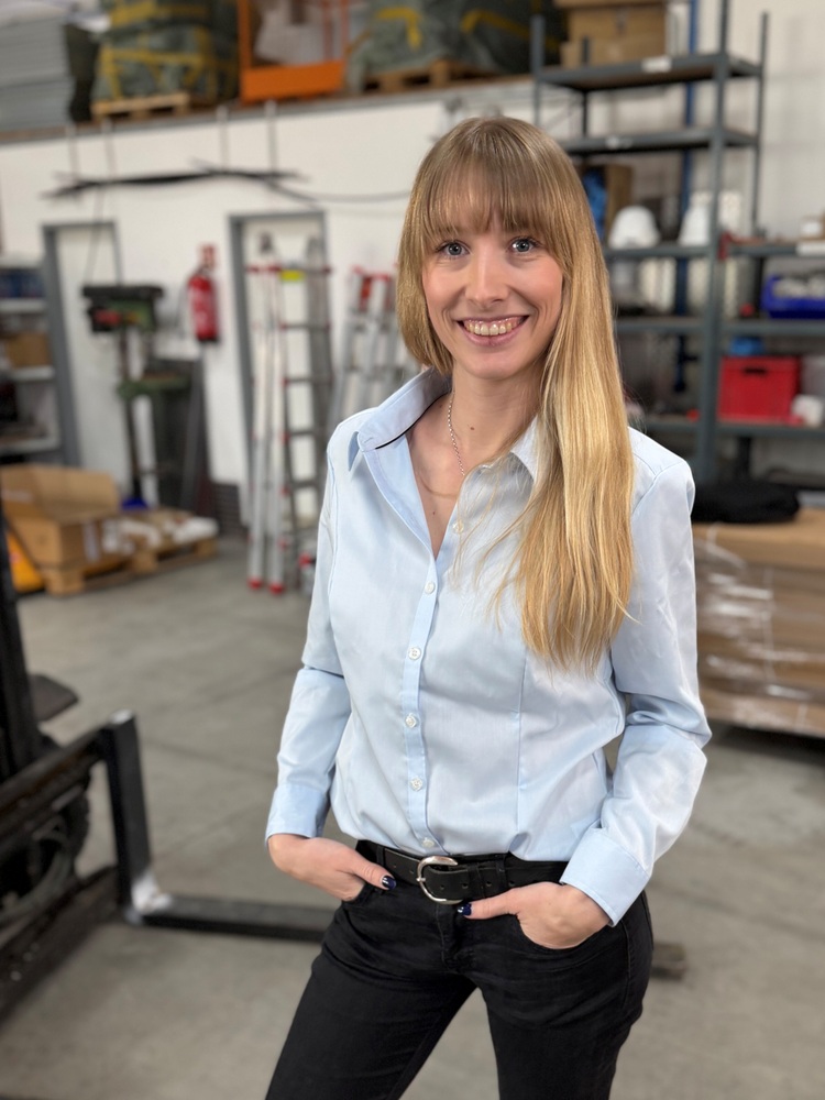 Ingenieurin mit langem blondem Haar lächelt in Lagerhalle, trägt hellblaues Hemd und schwarze Hose, Symbol für Frauen in der Gebäudetechnikbranche.