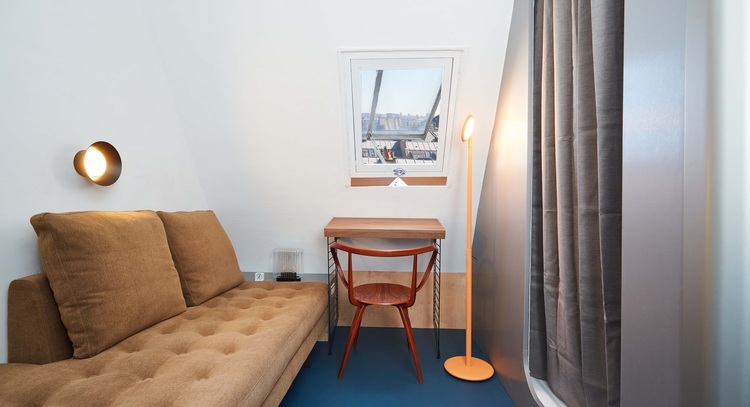 Kompaktes Arbeitszimmer mit braunem Sofa, Holzschreibtisch, Stehlampe, Wandbeleuchtung und Fenster mit Blick auf moderne Stadtarchitektur.