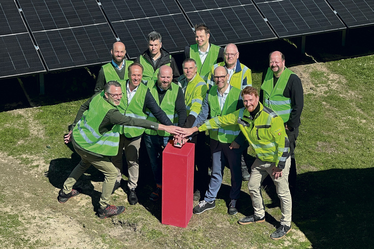 Fachpublikum in grünen Warnwesten diskutiert im Freien an rotem Stehpult vor PV-Anlage, Fokus auf Gebäudetechnik und erneuerbare Energien.