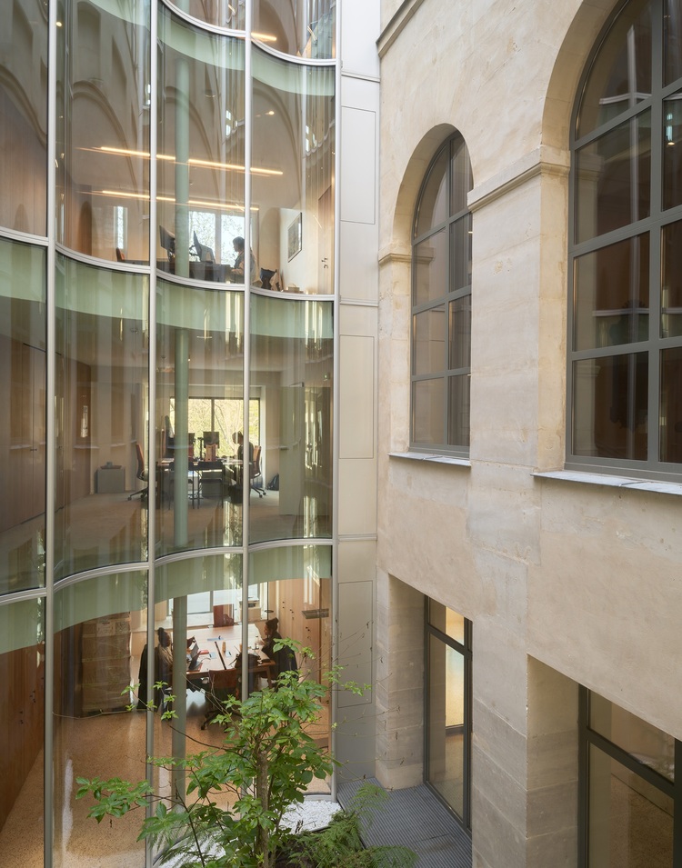Mehrgeschossige, moderne Bürolandschaft mit Glaswänden, neben historischen Steinwänden und großen Rundbogenfenstern in einem Bürogebäude.
