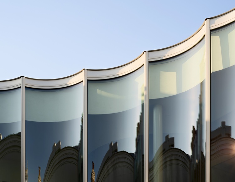 Gebogene Glasfassade eines modernen Bürogebäudes, spiegelt umliegende Architektur wider, Beispiel für energieeffiziente Gebäudehülle bei Sonnenschein.