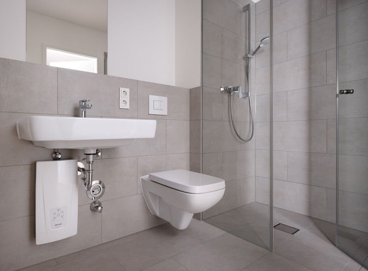 Barrierefreies Badezimmer mit wandhängendem WC, Waschtisch und bodengleicher Glasdusche auf hellgrauen Fliesen, geeignet für moderne Gebäudetechnik.