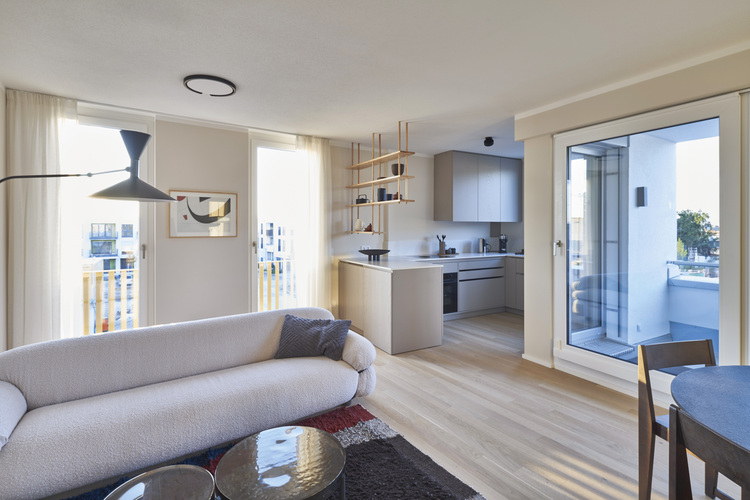 Heller Wohnbereich mit weißem Sofa, offener Küche und großen Fenstern - Beispiel für energieeffiziente Gebäudetechnik und Tageslichtnutzung.