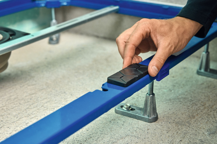 Eine Hand justiert ein schwarzes Kunststoff-Bauteil an einem blauen höhenverstellbaren Metallgestell für gebäudetechnische Installationen auf Beton.